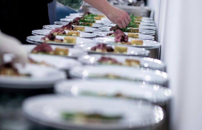Side view of male cook in apron serving many plates with delicious food while preparing lunch in restaurant at big table